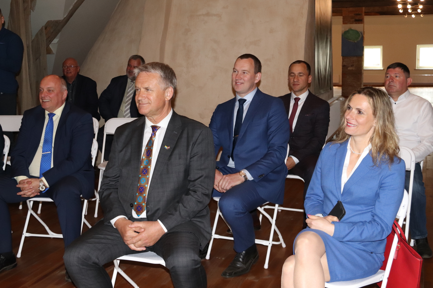 Viesi sēž krēslos un skatās prezentāciju